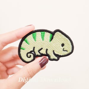 May include: A green and yellow embroidered iron-on patch of a cartoon chameleon. The chameleon is curled up with its tail wrapped around its body.