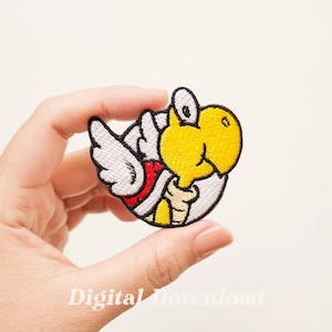 Pode incluir: Etiqueta bordada para passar a ferro com um Koopa Troopa amarelo com uma concha branca e detalhes vermelhos, a voar com asas brancas.