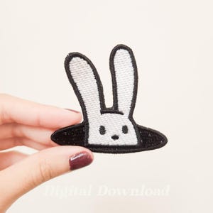 May include: White rabbit embroidered patch with black outline, peeking out of a black hole. The patch is perfect for adding a whimsical touch to clothing, bags, or other accessories.