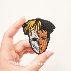 May include: Embroidered iron-on patch featuring a portrait of a person with half of their face as a skull. The person has black hair with yellow highlights and is wearing a black and white shirt.