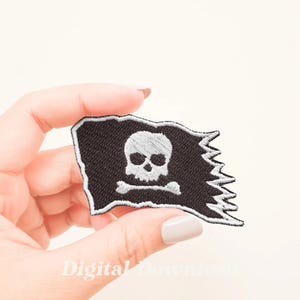 Puede incluir: Un parche de bandera pirata bordado en negro con un diseño de calavera y huesos cruzados blancos. La bandera está ligeramente deshilachada en los bordes.