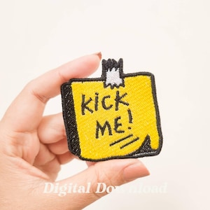 Puede incluir: Parche amarillo bordado con puntadas negras. El parche tiene forma de nota adhesiva con una esquina doblada y dice "Kick Me!" en texto negro.