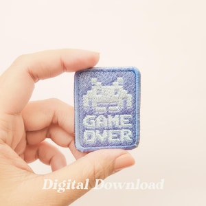 以下が含まれることがあります： 白いピクセル化されたエイリアンと「GAME OVER」のテキストが書かれた紫色の刺繍パッチです。