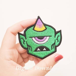 Peut inclure: Écusson brodé représentant un monstre cyclope vert avec un seul œil, une corne violette et jaune, et des détails noirs. L'écusson est tenu dans une main sur fond blanc. Le texte "Digital Download" est partiellement visible.