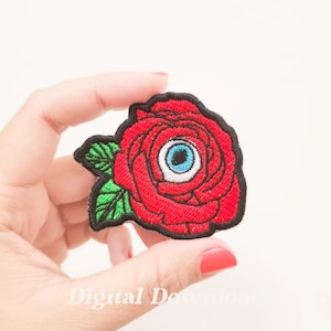 Puede incluir: Rosa roja bordada con un ojo azul en el centro. La rosa está sobre un fondo blanco.