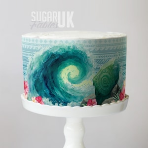May include: A round cake decorated with a blue and green wave design, a green stone, and pink flowers. The cake is presented on a white cake stand. The text "Sugar UK Fables" is visible on the top of the cake.