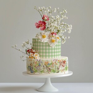 A two-tiered cake decorated with white flowers and greenery, placed on a white cake stand.