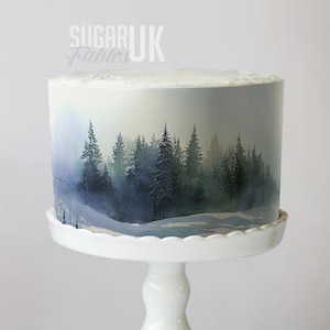 Op de afbeelding: Een ronde taart met een winterbosscène op de zijkant, gepresenteerd op een witte taartstandaard. De taart heeft een aquarelontwerp van groenblijvende bomen in blauwe en groene tinten, met de tekst "SUGAR Fables UK" bovenaan.