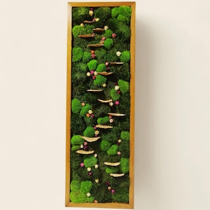 Może przedstawiać: Prostokątne dzieło sztuki ściennej w drewnianej ramie, wypełnione żywym zielonym mchem. Małe, jasno zabarwione drewniane platformy ułożone są pionowo, przeplatane maleńkimi kolorowymi kwiatami. Dzieło jest naturalnym, dekoracyjnym elementem.