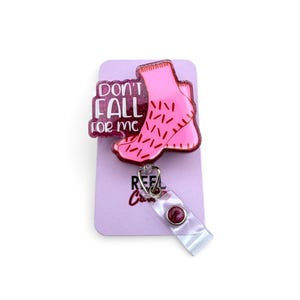 May include: A pink badge reel with the text "Don't Fall For Me" and a pink foot design with red accents. The badge reel has a clear plastic strap and a silver metal clip.