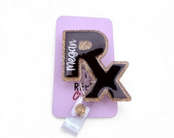 May include: A black and gold glitter badge reel with the letters "Rx" and the name "Megan" in white. The badge reel is attached to a purple card.