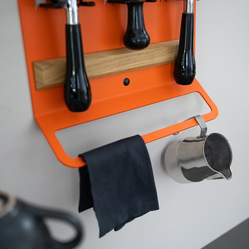 May include: An orange metal coffee station organizer with black handles and a wooden accent. A black cloth hangs from the bottom, and a stainless steel pitcher is attached to the side. The organizer is mounted on a white wall.