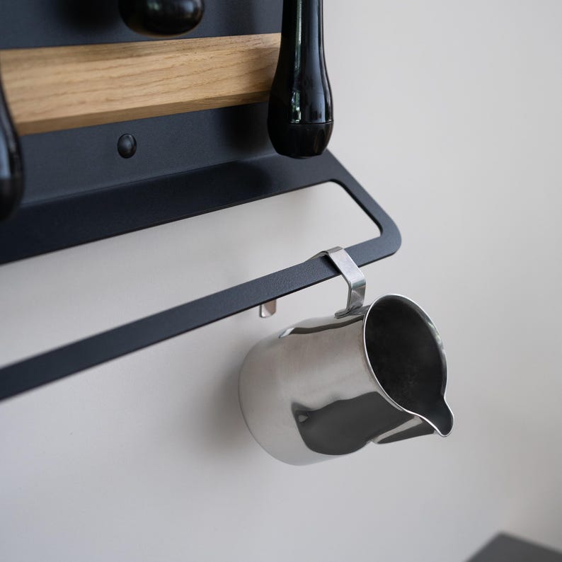 May include: A stainless steel milk frothing pitcher hangs from a black metal rack. The rack is mounted on a white wall, with a wooden shelf above it. Black utensils hang above the pitcher.