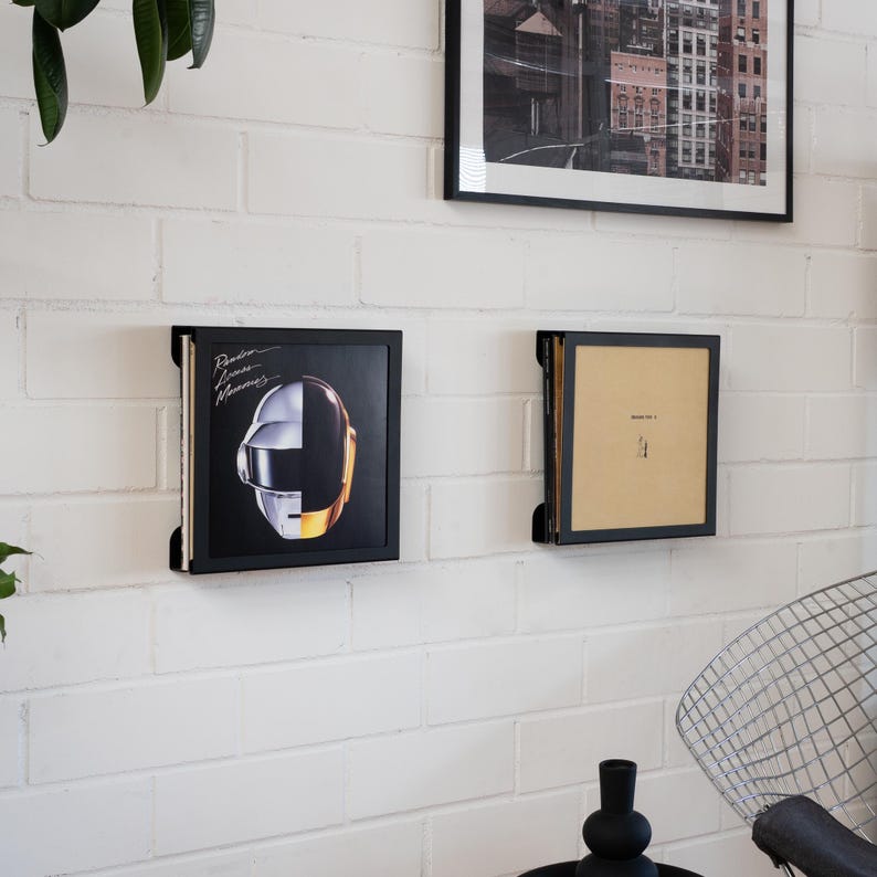 Pode incluir: Dois suportes pretos emoldurados para discos de vinil montados numa parede de tijolos brancos. Um exibe uma capa de &aacute;lbum Daft Punk com um design de capacete prateado e dourado. O outro cont&eacute;m um disco com uma capa bege.