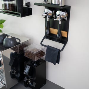 May include: Black metal wall-mounted coffee accessory rack with two chrome portafilters, a wooden shelf, and a black towel hanging below. The rack is mounted on a white wall next to a black coffee grinder with a clear bean hopper.