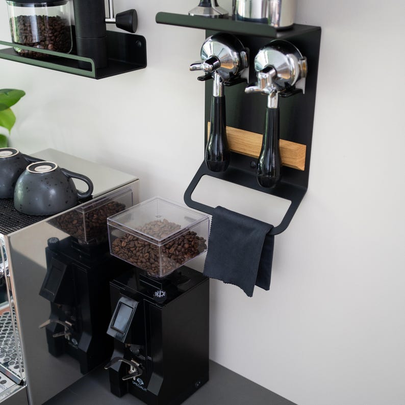 May include: Black metal wall-mounted coffee accessory rack with two chrome portafilters, a wooden shelf, and a black towel hanging below. The rack is mounted on a white wall next to a black coffee grinder with a clear bean hopper.