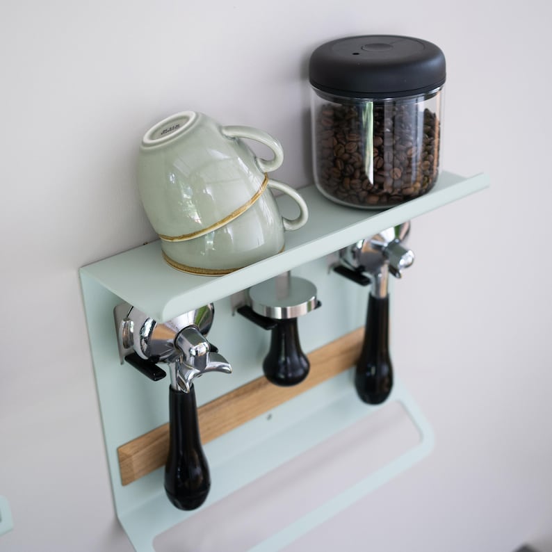 May include: A mint green coffee station wall shelf with two stacked light green mugs, a glass jar of coffee beans, and three espresso machine accessories. The shelf has a wooden accent and is mounted on a white wall.