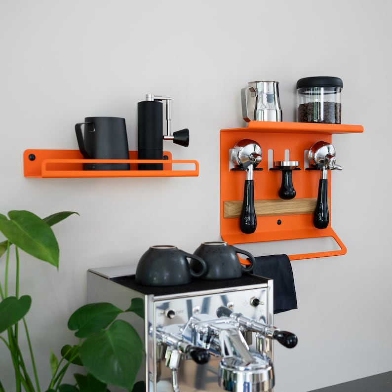 May include: Orange metal coffee station wall shelves with coffee accessories. Includes a coffee grinder, black mugs, espresso machine portafilters, a milk frothing pitcher, and a container of coffee beans. The shelves are mounted on a white wall.