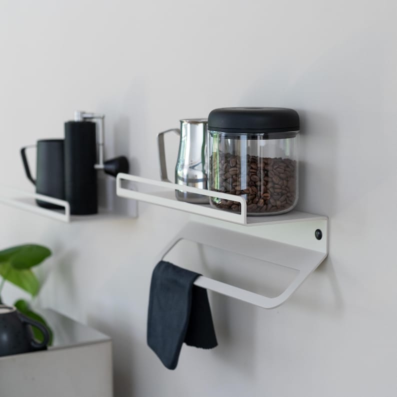 May include: Two white metal shelves mounted on a wall with a black towel rail below. The top shelf holds a black coffee grinder, a silver jug, and a glass jar with a black lid filled with coffee beans.