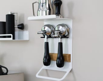 May include: White metal wall-mounted coffee station with two black and chrome portafilters, a stainless steel jug, and a black tamper. The station has a shelf and a towel rail.
