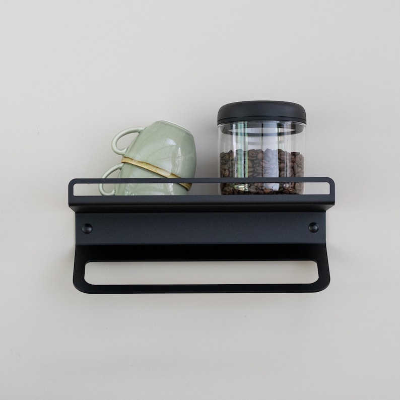 May include: Black metal wall shelf with a towel bar, a glass jar filled with coffee beans, and two green ceramic mugs.