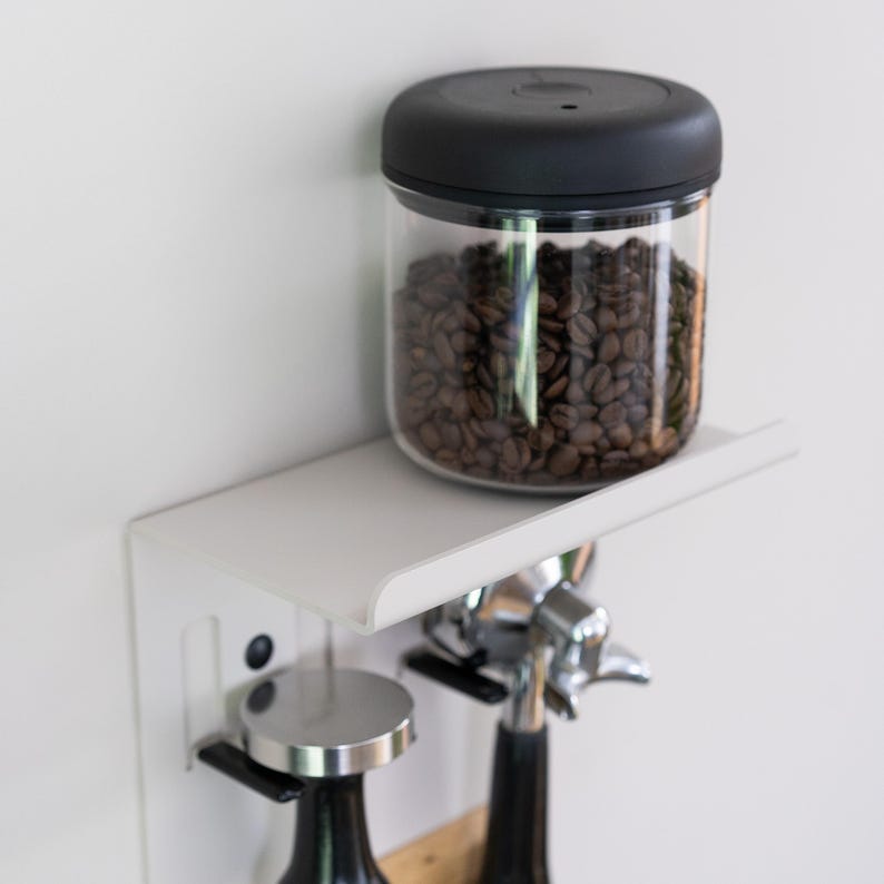 May include: A white wall-mounted shelf with a black lidded glass jar filled with coffee beans. Two silver and black espresso tampers hang below the shelf.