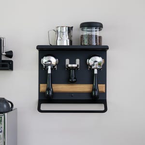 May include: Black metal espresso machine accessory organizer with a wooden accent. It holds two portafilters, a tamper, a stainless steel pitcher, and a glass jar with coffee beans. The organizer is mounted on a light gray wall.