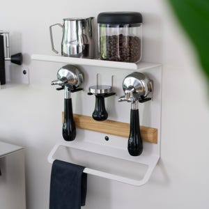 May include: A white wall-mounted coffee station with a shelf holding a stainless steel pitcher and a jar of coffee beans. Below, there are two espresso machine portafilters, a tamper, and a towel bar. The portafilters have black handles.