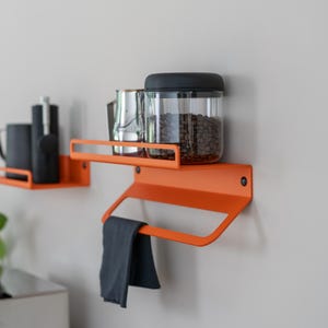 May include: Two orange metal wall shelves with a black tea towel hanging on the bottom shelf. The top shelf has a glass jar filled with coffee beans and a silver jug.