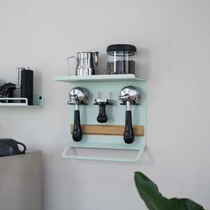 May include: A mint green coffee station organizer mounted on a wall. It features a shelf with a stainless steel pitcher and a glass jar with a black lid. Below, it holds two espresso machine portafilters, a tamper, and a wooden accent.