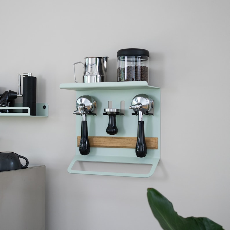 May include: A mint green coffee station organizer mounted on a wall. It features a shelf with a stainless steel pitcher and a glass jar with a black lid. Below, it holds two espresso machine portafilters, a tamper, and a wooden accent.