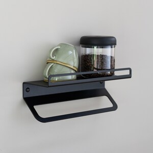 May include: Black metal wall shelf with a towel bar underneath. The shelf has a green ceramic mug and a glass jar with a black lid filled with coffee beans on top.