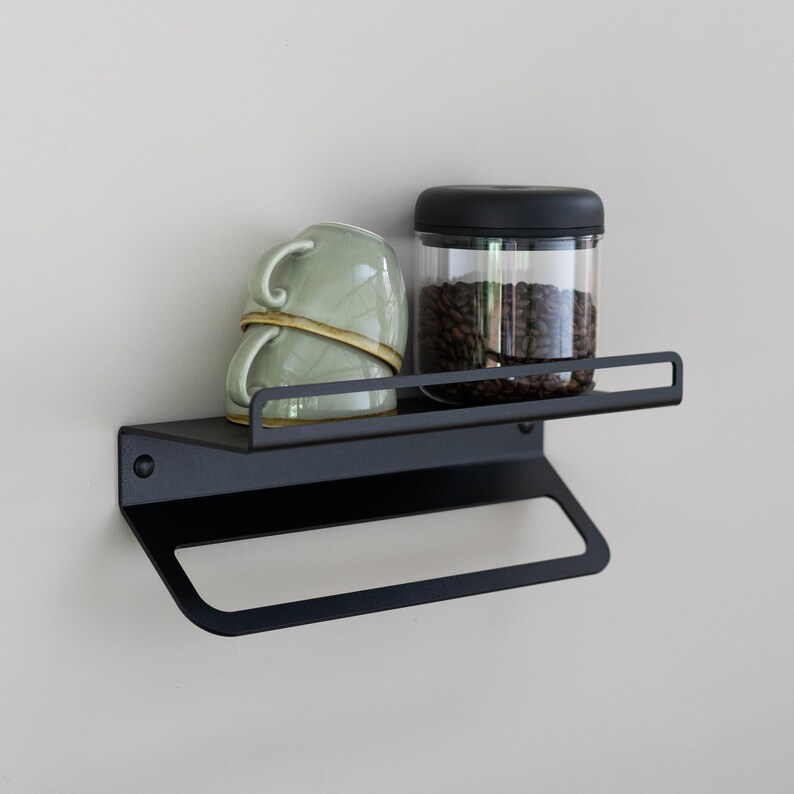 May include: Black metal wall shelf with a towel bar underneath. The shelf has a green ceramic mug and a glass jar with a black lid filled with coffee beans on top.