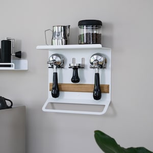 May include: White wall-mounted coffee station with a shelf holding a stainless steel pitcher and a jar of coffee beans. Below, two espresso machine portafilters with black handles and a tamper are mounted. A wooden accent strip adds warmth.