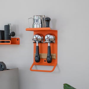 May include: Orange metal wall mounted coffee accessory organizer with two portafilter holders, a shelf, and a towel bar. A stainless steel milk pitcher and a black tamper are on the shelf.