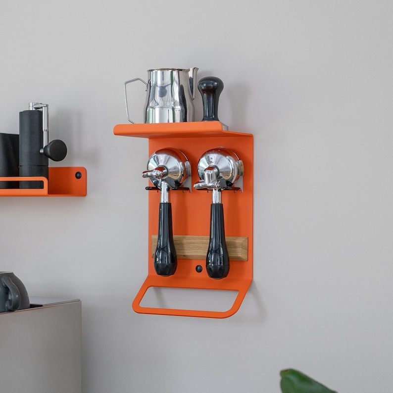 May include: Orange metal wall mounted coffee accessory organizer with two portafilter holders, a shelf, and a towel bar. A stainless steel milk pitcher and a black tamper are on the shelf.