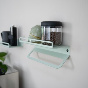 May include: Two light green metal shelves mounted on a white wall. The top shelf has a glass jar with a black lid filled with coffee beans, a white ceramic bowl, and a small black bottle. The bottom shelf has a towel bar.