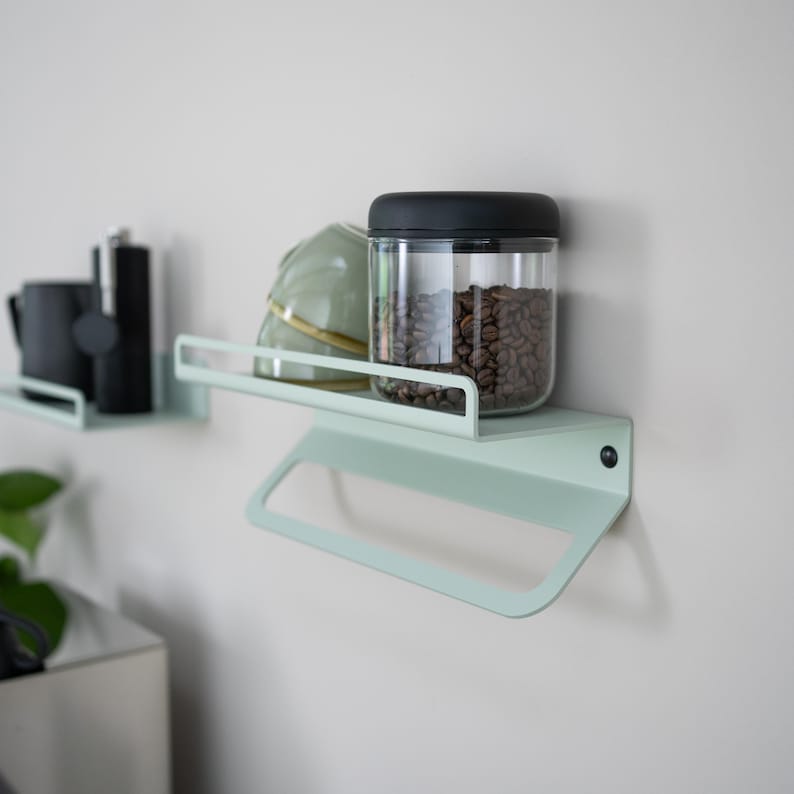 May include: Two light green metal shelves mounted on a white wall. The top shelf has a glass jar with a black lid filled with coffee beans, a white ceramic bowl, and a small black bottle. The bottom shelf has a towel bar.