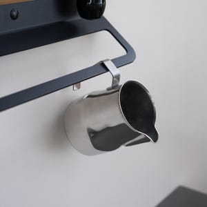 May include: A stainless steel milk frothing pitcher hanging from a black metal rack. The pitcher has a curved handle and a spout.