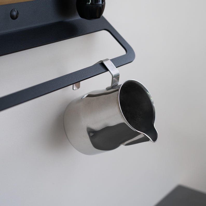 May include: A stainless steel milk frothing pitcher hanging from a black metal rack. The pitcher has a curved handle and a spout.