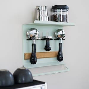 May include: A mint green coffee station wall organizer with a shelf holding a stainless steel pitcher and a jar of coffee beans. Below are two chrome portafilters with black handles, a tamper, and a wooden accent.