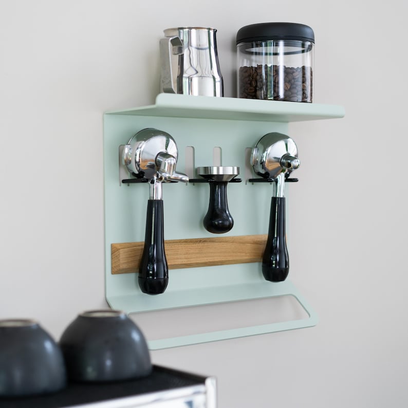 May include: A mint green coffee station wall organizer with a shelf holding a stainless steel pitcher and a jar of coffee beans. Below are two chrome portafilters with black handles, a tamper, and a wooden accent.