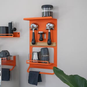 Op de afbeelding: Een oranje metalen wandkoffiestation met planken, met espressomachine portafilters, een stamper, een glazen pot met koffiebonen en een roestvrijstalen mok. Een donkergrijze handdoek hangt eronder.