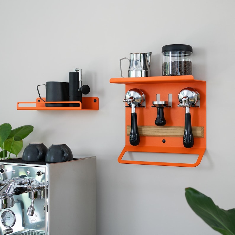 May include: Orange metal wall-mounted coffee station with a shelf holding a black mug and grinder. Another station features espresso machine portafilters, a tamper, and a jar of coffee beans. A stainless steel espresso machine is in the foreground.