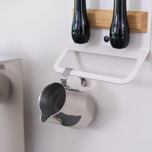 May include: A white metal kitchen organizer with two black metal hooks holding a pair of black metal whisks. A stainless steel milk frother hangs from a hook on the bottom of the organizer.