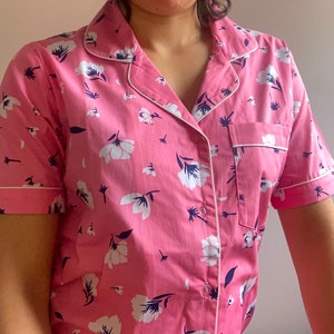 May include: Pink pajama set with a white floral pattern and navy blue accents. The short-sleeved top has a chest pocket and white piping along the edges. The fabric is a soft, lightweight material.
