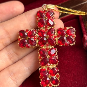 May include: A gold-coloured cross pendant necklace featuring many oval, deep red gemstones. The cross is set with smaller red stones, creating a sparkling, ornate design. The necklace is displayed against a red velvet background.