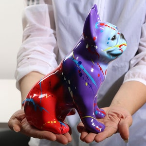 Peut inclure: Une figurine en céramique peinte à la main d'un bouledogue français. Le chien est assis, la tête tournée sur le côté, et son corps est peint dans des tons de rouge, violet, bleu et jaune.