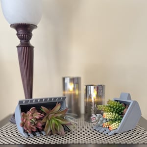 Puede incluir: Dos cubos de excavadora en miniatura de plástico gris llenos de suculentas artificiales. Los cubos están sobre un mantel tejido marrón.