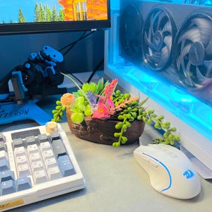 May include: A small, decorative planter filled with colorful faux succulents and flowers. The brown planter sits on a desk next to a white keyboard and mouse. A computer monitor and a computer tower are in the background.